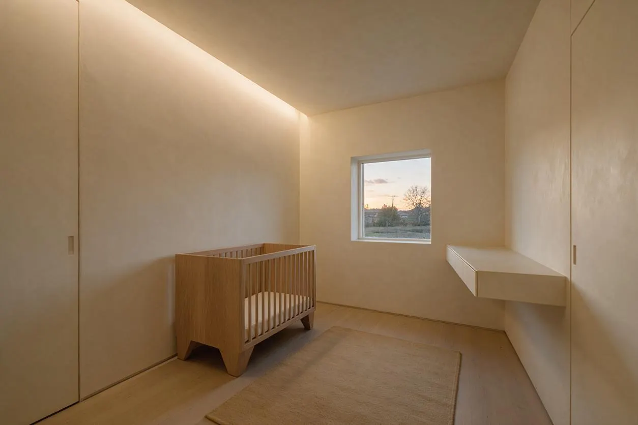 A sparse minimalist nursery with smooth beige walls, a natural oak crib with vertical slats, and a neutral rug on the floor. A small window brings in soft sunset light, and a floating wall-mounted shelf adds a simple functional detail.