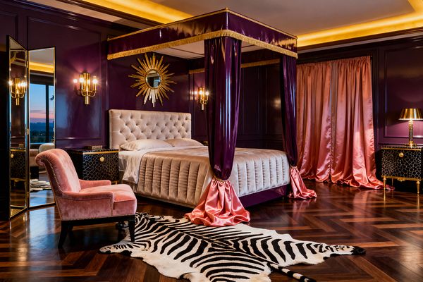 A glamorous bedroom with deep aubergine lacquered wall paneling and a four-poster canopy bed draped in glossy purple and blush satin with gold fringe. The tufted champagne velvet headboard is flanked by brass sconces and black reptile-textured nightstands; a gilded sunburst mirror, pink velvet slipper chair, dark herringbone wood floors, zebra hide rug, mirrored screen, and warm cove lighting complete the Hollywood Regency scene.