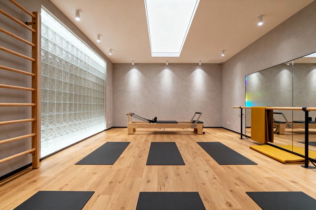 A minimalist home gym with light oak flooring features evenly spaced black mats facing a central wood Pilates reformer. The space has soft gray walls, a backlit glass-block wall with adjacent wooden stall bars, a full-width mirror and wood-and-black metal barre on the right, and a large rectangular skylight with recessed spotlights providing diffuse, even lighting, creating a calm, bright environment for stretching, barre, and Pilates work.