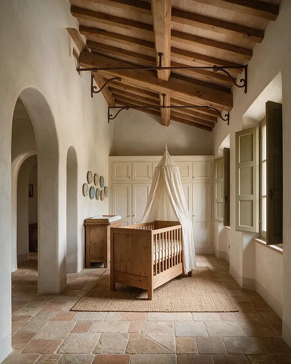 A Mediterranean-style nursery with a wooden crib draped in a soft white canopy, set on a natural jute rug over terracotta floor tiles. The room features arched plaster openings, cream built-in cabinetry, olive-green window shutters, and exposed timber سقiling beams that create a warm, serene atmosphere.