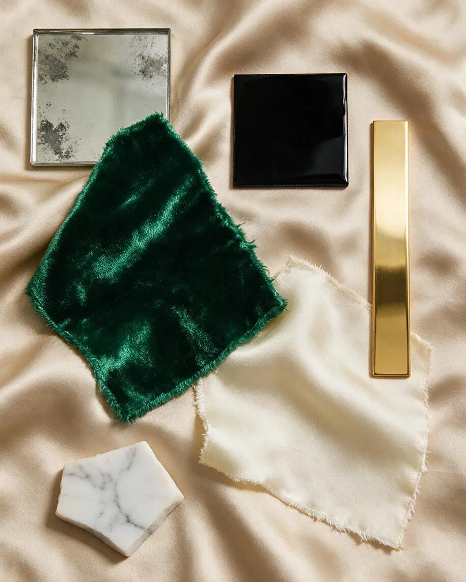 A styled material palette is arranged on shimmering champagne satin, featuring a square emerald green velvet swatch, a glossy black tile, a mirrored silver tile, a slim polished gold bar, and a white marble sample. The mix of luxe textures and reflective finishes creates a classic Hollywood Regency look.