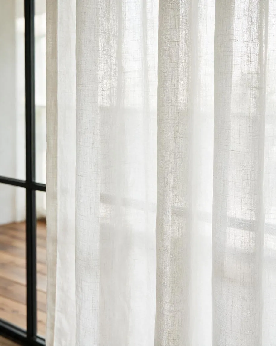 A close-up of sheer white linen curtains hanging beside a black metal-framed window. Soft daylight filters through the lightweight fabric, with warm wood flooring faintly visible in the background.