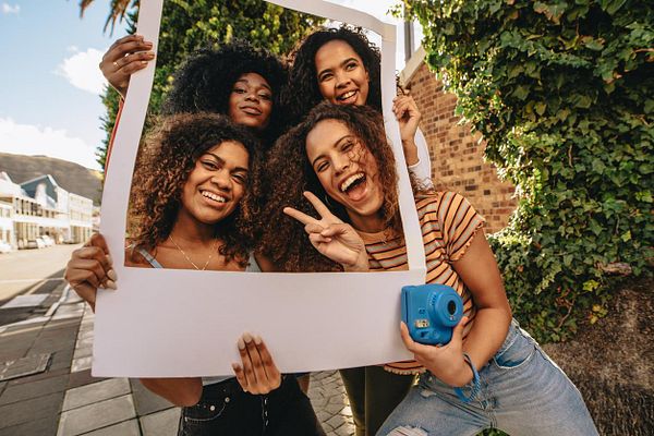 Four women are joyfully posing together with a large frame in a vibrant outdoor setting.