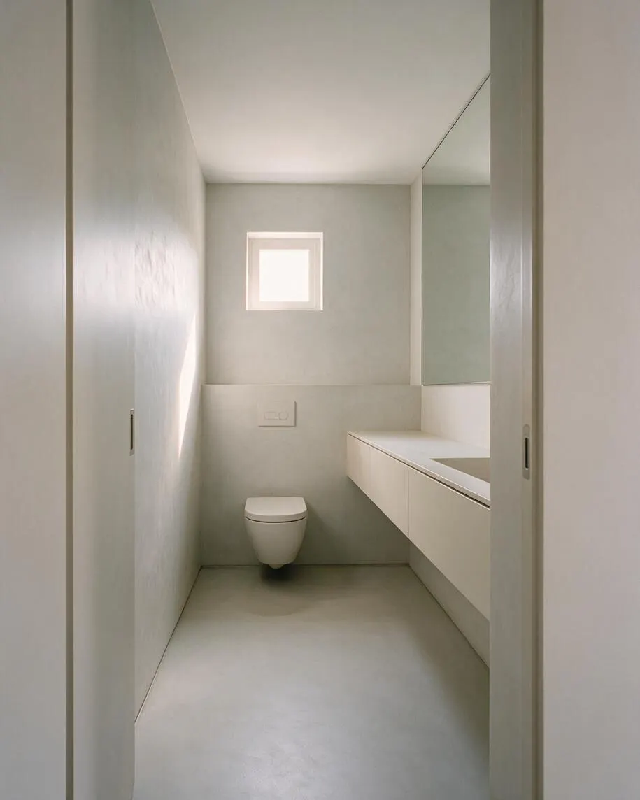 A narrow minimalist powder room with pale gray walls, a small square frosted window at the back, and a wall-hung toilet centered below it. On the right, a long floating vanity with a white counter and integrated sink extends toward the camera, while a large mirror above reflects light and enlarges the compact space.