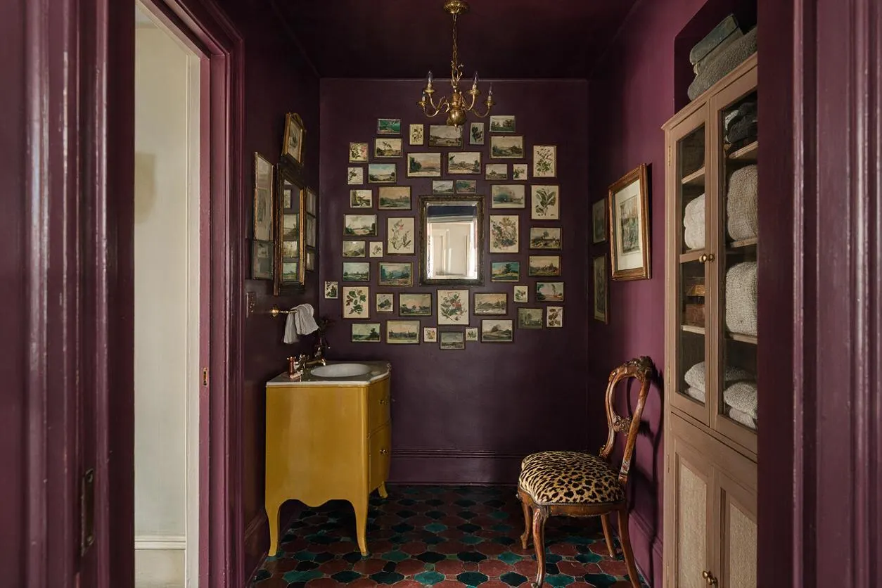 A compact powder room with deep plum walls and ceiling, a small yellow vanity with a round sink, and a brass chandelier overhead. The back wall is filled with a dense gallery of framed botanical and landscape prints, while a leopard-print chair and built-in cabinet with folded towels add layered, maximalist detail.