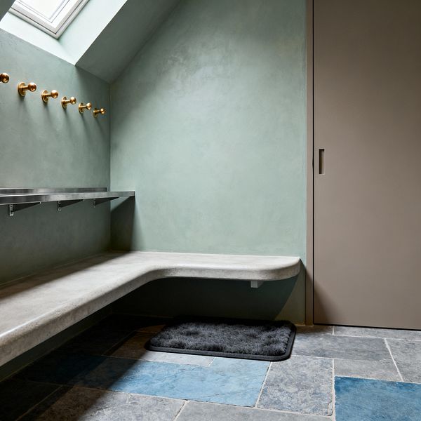 A minimalist mudroom with sage green plaster walls and a sloped ceiling lit by a skylight. An L-shaped floating concrete bench lines two walls, paired with a slim stainless-steel shelf and a row of polished brass hooks. A taupe sliding door with a recessed pull is on the right. The floor is paved in large blue-gray slate tiles, with a small charcoal mat beneath the bench.