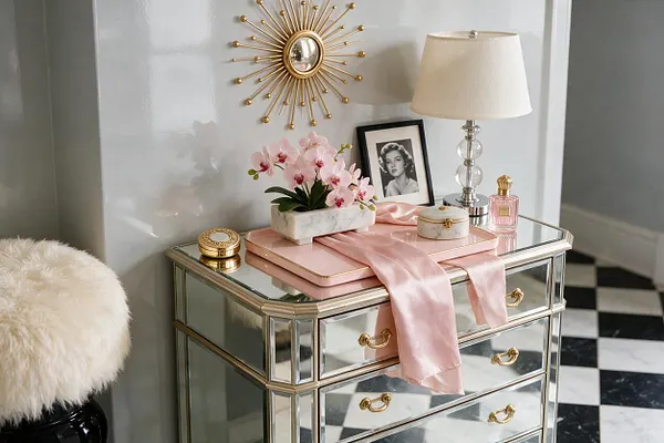 A Hollywood Regency-inspired vignette features a mirrored console with gold trim and drawer pulls, styled with a blush tray, satin ribbon, pink orchids, a framed black-and-white portrait, a clear glass lamp with a cream shade, and a sunburst wall mirror. Black-and-white checkered flooring and a faux fur stool add to the glamorous look.