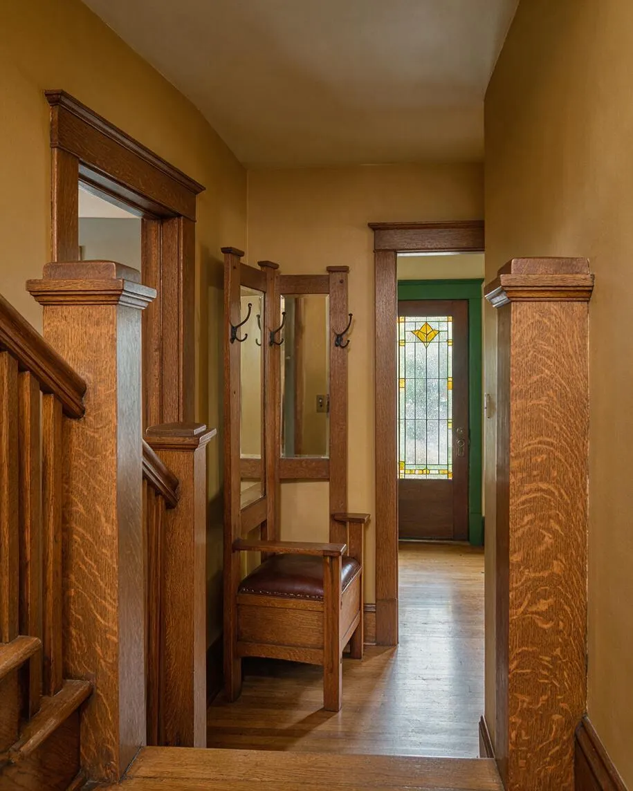 A narrow Craftsman-style entryway with rich wood trim, thick square newel posts, and a built-in bench with dark leather seating. The hall is painted in warm golden tones and leads to a front door with decorative stained glass, creating a cozy, historic feel.