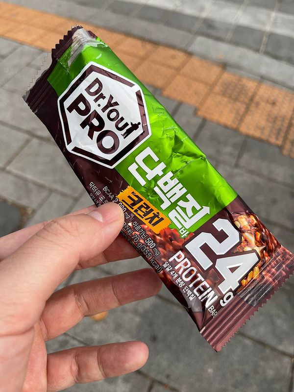 A hand holds a protein bar wrapper against a textured pavement background.