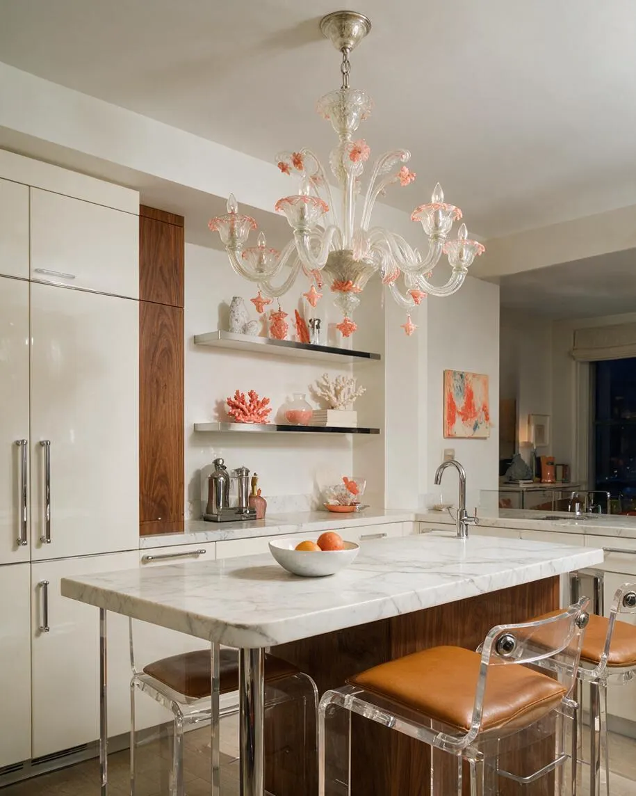 A bright eclectic kitchen features a white marble island with a waterfall edge, clear acrylic bar stools with tan seats, and sleek white cabinetry accented by vertical wood panels. Above the island hangs a clear glass chandelier with pink floral details, while open shelves display coral-toned decor and art adds color to the wall.