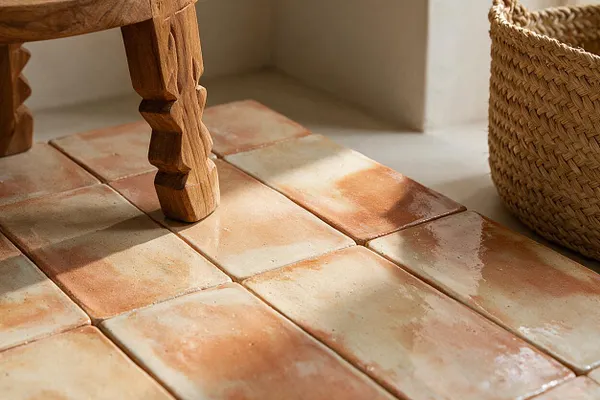 A close-up view of handmade terracotta floor tiles in warm cream and burnt orange tones, with a carved wooden furniture leg and a woven basket nearby. Sunlight casts soft shadows across the textured surface, creating a cozy bohemian feel.
