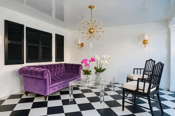 A stylish backyard lounge with a black-and-white checkerboard floor, white walls, and black window shutters. The space features a plush purple velvet tufted sofa, black wooden chairs with cream cushions, a clear acrylic coffee table, mirrored floral planters, and a gold starburst chandelier overhead.