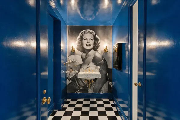 A narrow Hollywood Regency powder room with glossy cobalt-blue walls, a black-and-white checkerboard floor, and a large black-and-white portrait mural at the far end. A marble sink with gold fixtures and brass sconces adds glamour, while mirrored and polished surfaces reflect light.