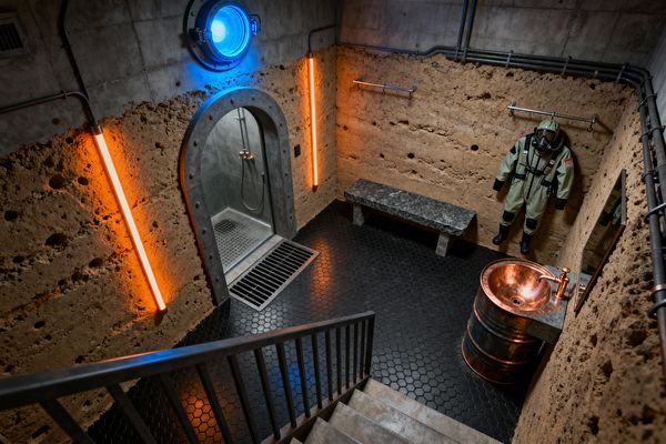 A subterranean mudroom features rough rammed-earth walls topped with concrete, black hex tile flooring, a riveted steel-framed decontamination shower beside a floor drain, a stone bench under wall hooks holding a hazmat-style suit, and a copper barrel sink with matching faucet below a simple mirror. A blue porthole light and two vertical orange neon tubes cast moody industrial illumination across exposed conduit and the stair rail.