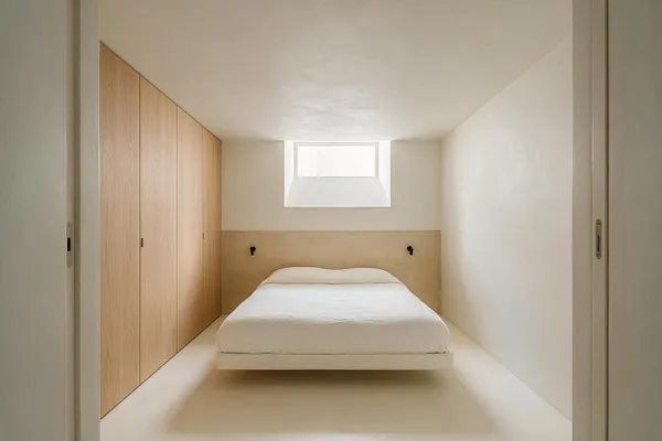 A compact minimalist basement bedroom with a floating bed centered against a simple beige headboard wall, framed by white walls and a small high window. Full-height light oak built-in wardrobes line the left side, while the room stays uncluttered with soft neutral finishes and clean lines.
