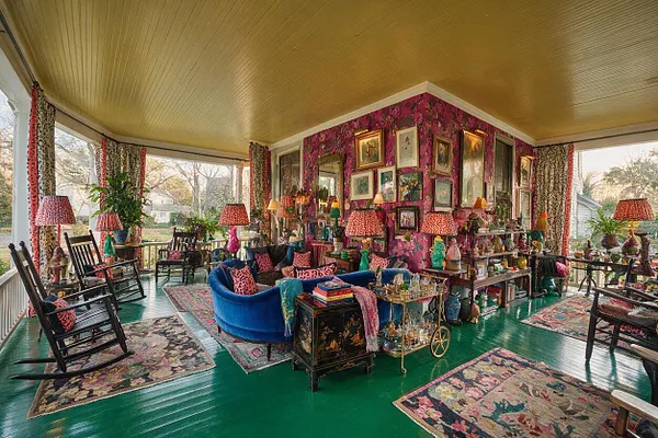 A covered front porch transformed into a colorful maximalist sitting room with a glossy green floor, golden ceiling, and floral wallpaper on the central wall. The space is filled with mismatched vintage chairs, a bright blue sofa, layered patterned rugs, framed artwork, table lamps, and decorative objects clustered across shelves and tables.
