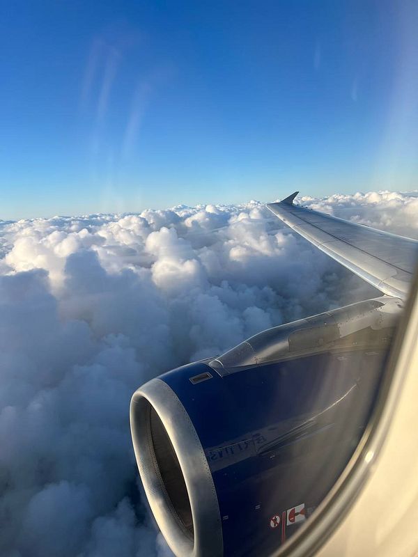 A view from an airplane window showcasing the wing and engine above a sea of clouds under a clear blue sky.