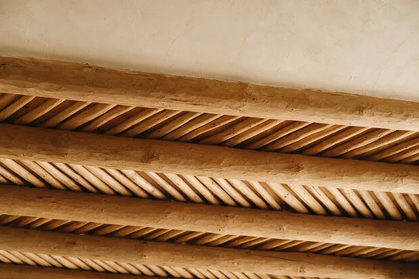 An angled view looking up at a ceiling with thick rustic wood beams and narrow diagonal wood slats between them. The warm honey-brown wood contrasts with a smooth pale plaster ceiling, creating a Southwestern-inspired architectural detail.
