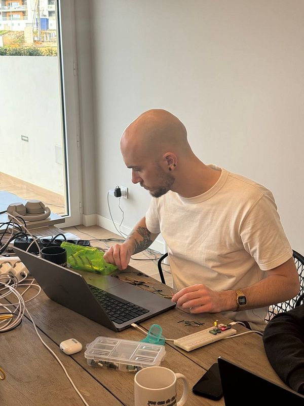 A man is working at a coworking space, focused on a laptop while handling electronic components.