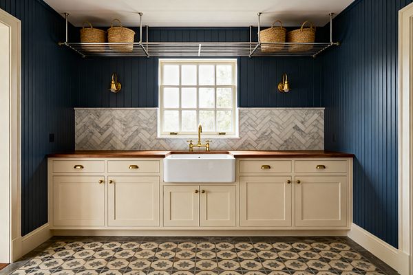 Traditional laundry room with a centered white farmhouse sink beneath a divided-light window, flanked by cream Shaker cabinets with brass cup pulls and knobs and warm wood countertops. Deep navy vertical beadboard, a Carrara marble herringbone backsplash, brass bridge faucet and sconces, patterned gray-beige floor tiles, and a ceiling-mounted stainless drying rack with woven baskets complete the symmetrical composition.