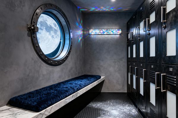 A futuristic mudroom with charcoal gray plaster walls features a round, riveted porthole-style window showing a large moon image. A cantilevered marble bench with a deep navy plush cushion faces matte black metal lockers with frosted panels and long stainless pulls. A crystal bar light throws rainbow reflections across the wall, while black raised-dot rubber flooring catches scattered silver glitter near the entrance.