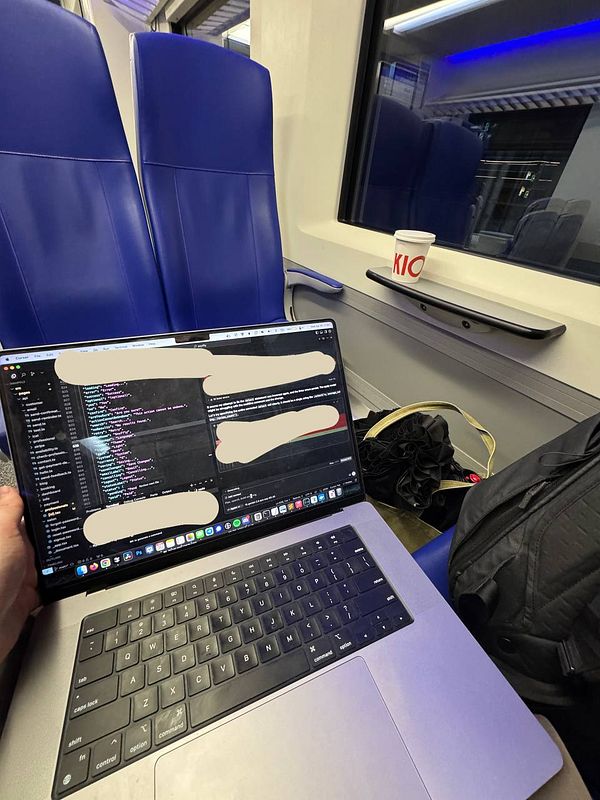 A person is working on a laptop while sitting in a train, surrounded by blue seats and a coffee cup.