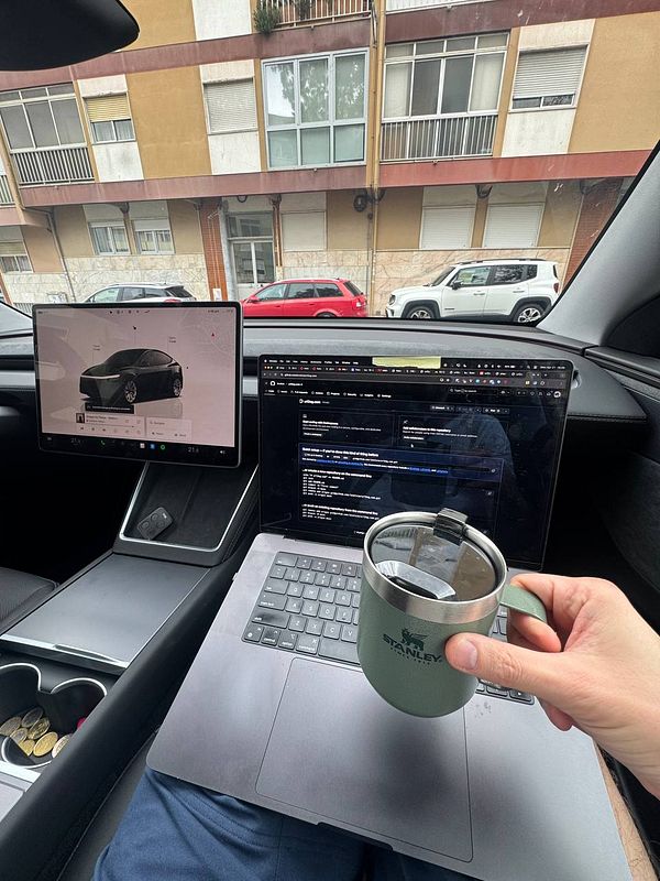 A person is working inside a Tesla Model Y, using a laptop and a mobile coffee cup.