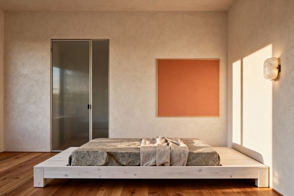 Sunlit minimalist kids’ room with a low whitewashed pine platform bed topped by a stone‑like gray cushion and a draped linen throw. Warm white plaster walls frame a large terracotta cork board and a ribbed frosted‑glass sliding door; a brass‑mounted frosted sconce glows on the right above wide‑plank natural wood flooring, with long window shadows adding calm in an uncluttered, ground‑level layout.