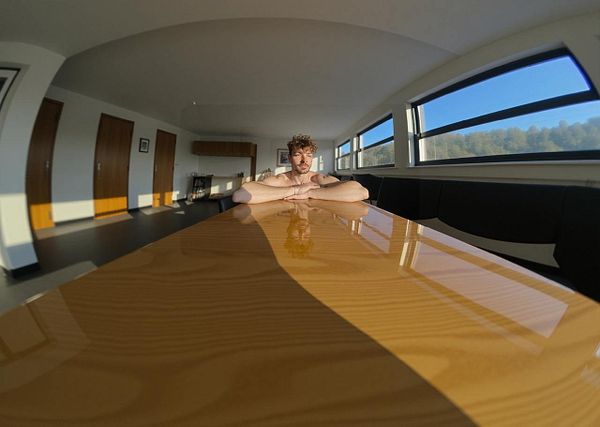 A person sits at a long wooden table in a well-lit room with large windows.