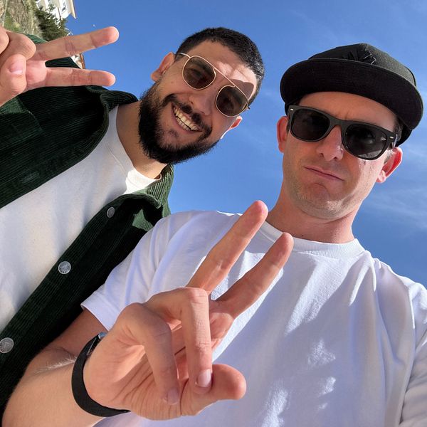 Two men pose for a selfie outdoors, both making peace signs with their hands.