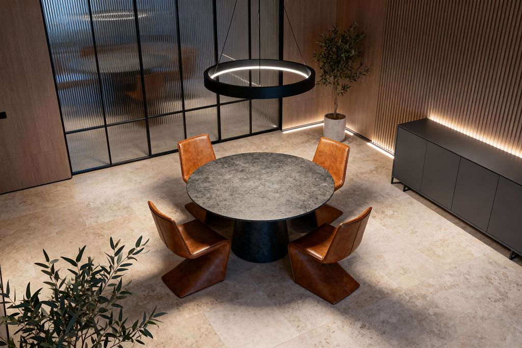 An overhead view of a modern dining room shows a round gray stone table on a black pedestal surrounded by four cognac leather cantilever chairs atop large beige stone tiles. A black circular LED pendant hangs above; a slatted light-wood wall and matte black sideboard are backlit by concealed LEDs, a reeded glass partition with black frames divides the space, and a potted indoor tree in a concrete planter completes the minimalist setting.