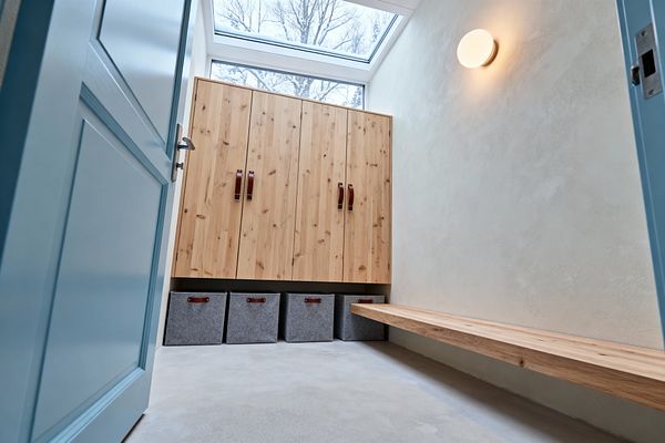 Compact Scandinavian mudroom with a large skylight above tall natural wood cabinets fitted with leather loop handles. Four gray felt bins line up beneath the cabinetry. A long floating wood bench runs along the right wall under a warm round wall sconce. The walls are textured warm white, the floor is light gray concrete, and a muted blue door with a metal handle opens into the bright, minimalist space.