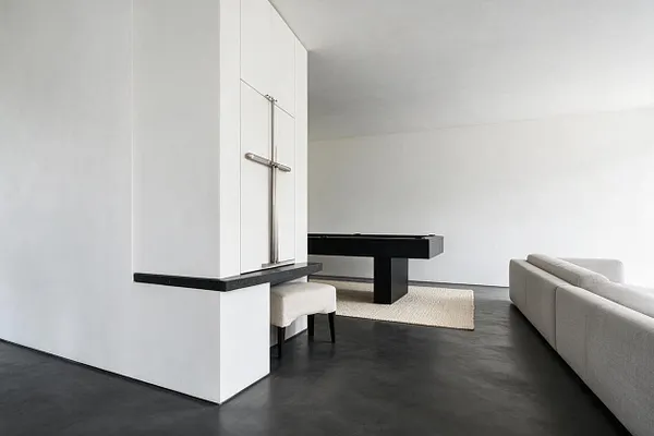 A minimalist game room with white walls, a dark polished floor, and a black pool table centered on a light woven rug. A cream upholstered sofa sits along the right side, while a small upholstered stool and a built-in white column with a metal cross-like detail add sculptural simplicity.