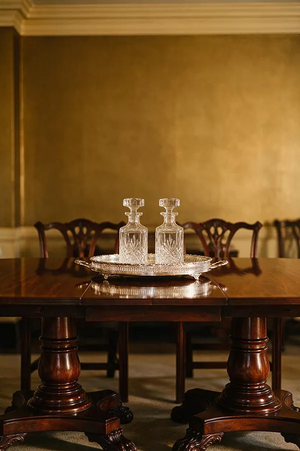 A formal Traditional dining room scene with a polished dark wood table, turned pedestal legs, and carved wooden chairs in the background. On the table sits a silver tray holding two cut-glass decanters, set against warm gold walls and classic crown molding.