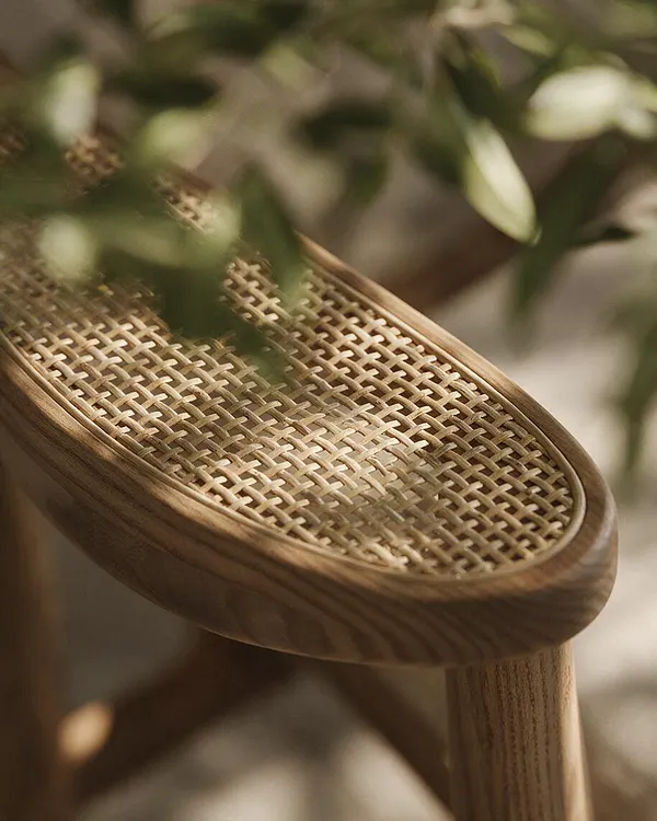 A close-up of a chair with a smooth curved wooden frame and a handwoven cane seat in a light natural finish. Soft green leaves blur in the foreground, reinforcing the biophilic feel and emphasizing organic materials and gentle light.