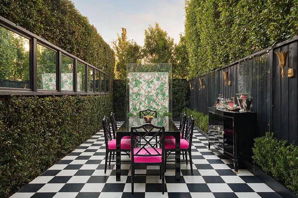 A narrow backyard dining area with a bold black-and-white checkerboard floor, surrounded by tall hedges and dark fencing. A black glass dining table with black chairs and bright pink cushions sits in the center, while a black sideboard and brass wall sconces line the right side, creating a polished Hollywood Regency look.