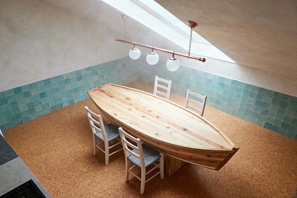 A coastal dining room beneath a sloped skylight features a boat-shaped natural pine table surrounded by four white ladder-back chairs with woven gray-blue seats. Sea-glass green square ceramic tiles line the lower wall, meeting warm white plaster above; a copper pipe pendant with three opal globes hangs overhead on chains, and the floor is cork.