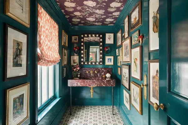 A narrow powder room with deep teal paneled walls and a matching teal trim, filled with framed artwork on both sides. A red marble wall-mounted sink with brass fixtures sits beneath a mirrored medicine cabinet, while a purple floral ceiling, patterned floor tiles, and a pink patterned window shade add bold maximalist detail.