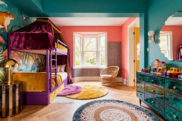 A vibrant children’s bedroom with teal walls and ceiling and a coral bay window wall. A purple bunk bed has a plum velvet canopy and mustard bedding against a tropical animal mural. The floor is herringbone oak with layered round rugs in gold, pink, cream, and blue. A rattan chair sits by the window; a glossy green dresser with brass knobs and a wavy mirror holds toy buildings. A striped side table with a brass dome lamp and a globe wall sconce add accents.