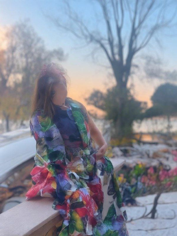 A woman in a colorful floral shawl sits on a ledge by a body of water during sunset.