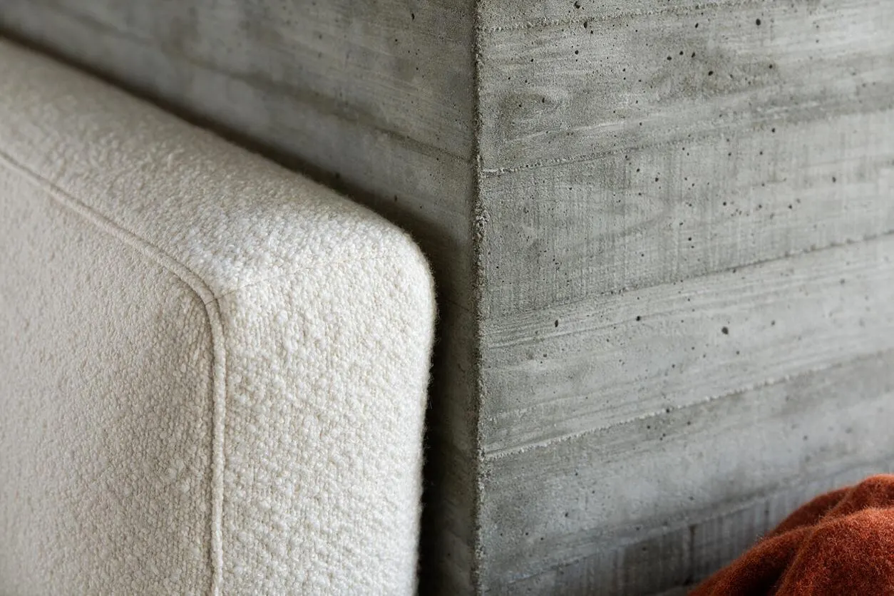 A close-up interior scene showing a cream bouclé upholstered seat beside a raw cast-concrete wall with visible formwork lines and small air pockets. A rust-colored textile peeks in at the lower right, adding warmth to the otherwise minimal, industrial composition.