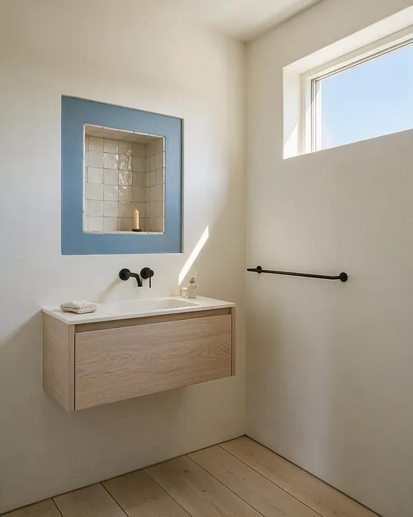 A bright Scandinavian-style powder room with a floating light oak vanity, white integrated sink, and matte black faucet. A recessed niche with blue trim and glossy white tile holds a single candle, while a small high window brings in natural light above a black towel bar and pale wood plank floor.