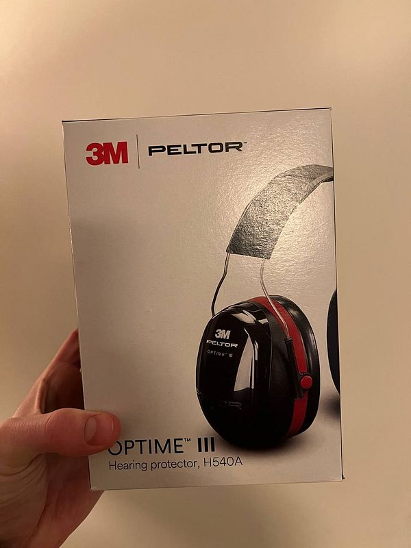 A person is holding a box of 3M Peltor Optime III hearing protectors.
