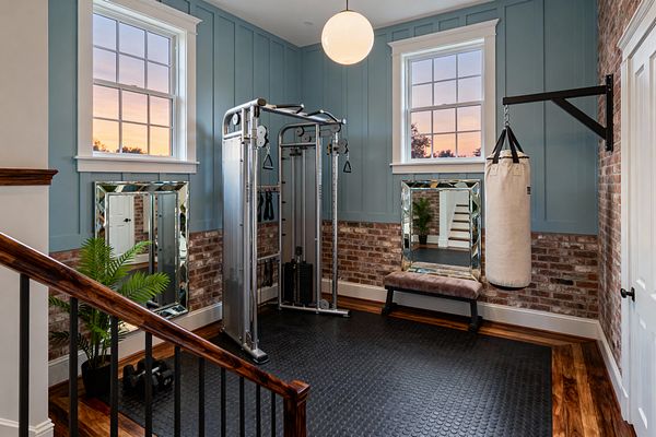 A compact home gym with dusty blue board-and-batten walls above a red-brick wainscot has two white-trimmed windows and a white globe pendant overhead. A stainless cable machine stands on black coin rubber flooring bordered by hardwood, with a cream heavy bag on a black wall mount, beveled mirrors leaning on the walls, a taupe velvet bench, a potted fern, and a stair rail with dark wood and black balusters in the foreground.