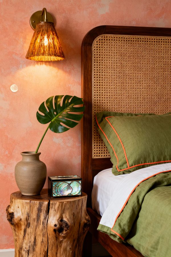A tropical bedroom vignette featuring a walnut bed with woven cane headboard, dressed in olive linen bedding with orange piping, against a mottled coral stucco wall. A brass wall sconce with a rattan shade glows warmly above a live-edge stump nightstand holding a clay vase with a single monstera leaf and a small abalone-inlaid box; white sheets peek beneath the duvet, creating a fresh contrast that emphasizes natural textures and earthy tones.