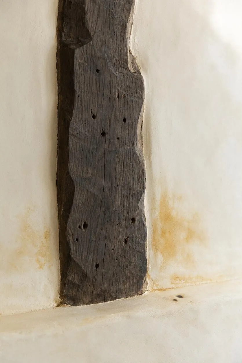 A close-up of a dark, weathered wooden beam set into a cream plaster wall. The rough wood grain, small knot holes, and aged patina contrast with the soft, warm-toned plaster surrounding it.