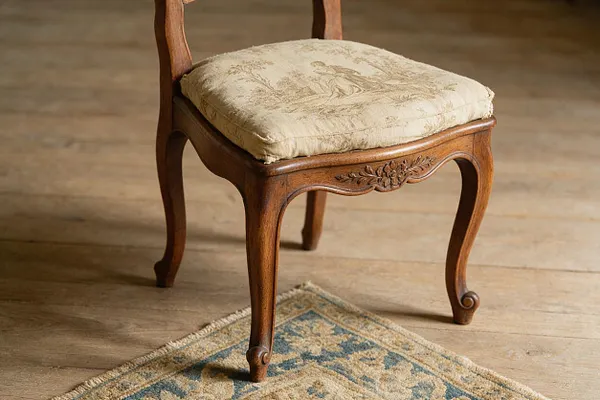 A vintage French Country chair with a carved wooden frame and curved cabriole legs, upholstered in a pale toile-pattern cushion. It sits on a light wood floor beside a muted blue-and-beige rug, with warm natural tones throughout.