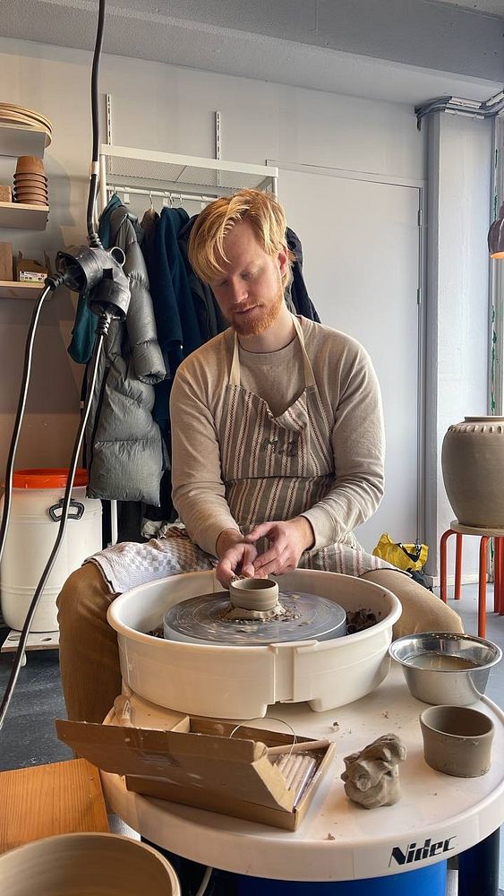 A close-up photograph of a hand holding a pottery mug filled with a creamy beverage and topped with a heart-shaped foam design.
