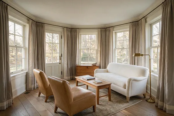 A bright transitional sunroom with many tall windows, beige drapery, and a curved wall of light-filled glass. The room includes a white sofa, two tan leather lounge chairs, a simple wood coffee table, a vintage wood chest, and a brass floor lamp on a pale patterned rug.