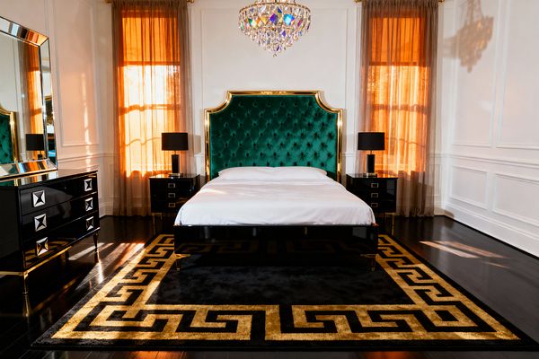 A Hollywood Regency bedroom centers on an emerald green, tufted velvet headboard framed in gold, flanked by glossy black lacquer nightstands topped with black-shaded lamps. A crystal chandelier hangs above white bedding and a black lacquer bed, with a black rug featuring a gold Greek key border on dark hardwood floors; white paneled walls and sheer taupe curtains glowing amber complete the glamorous scene.